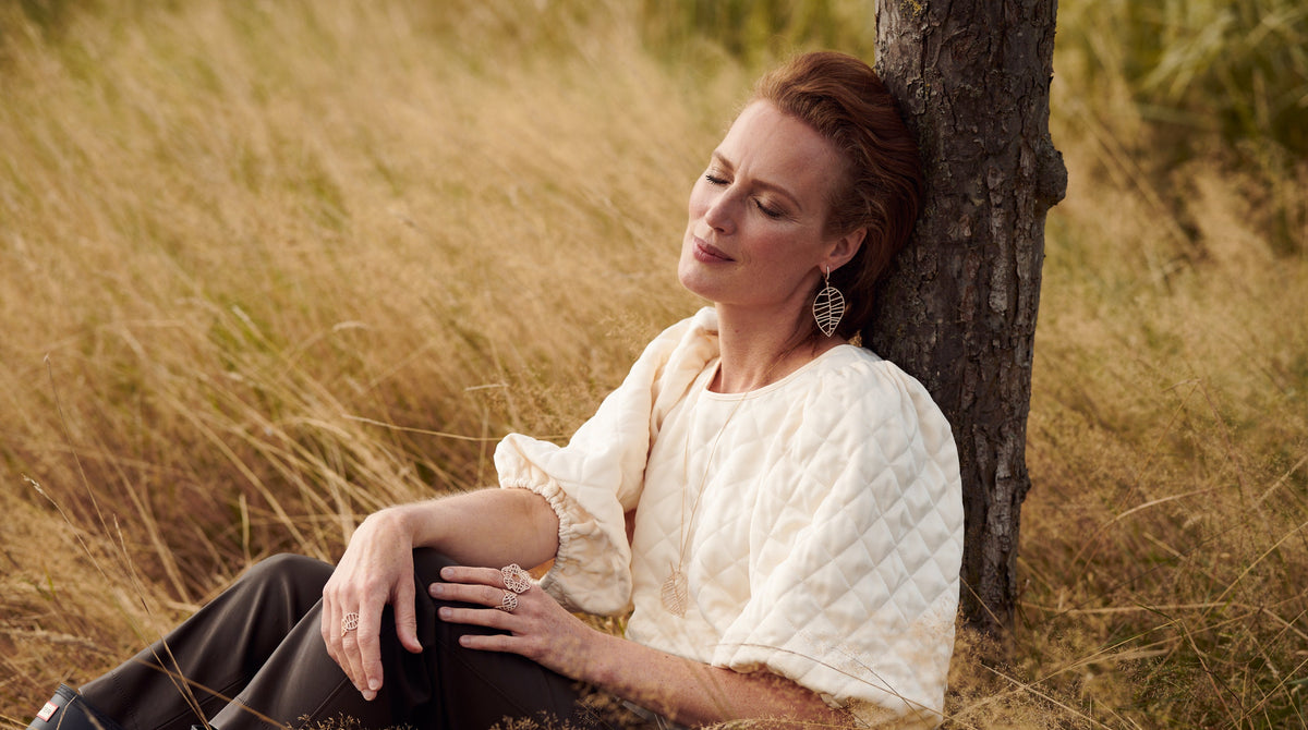 A woman with reddish-brown hair sits in a grassy field, leaning against a tree trunk. She is wearing 18k rose gold dangling earrings and a diamond set rings.