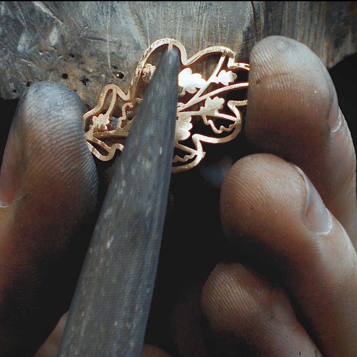 Close-up of a goldsmith's hands holding a 18k golden Oak pendant while using a metalworking tool, with a dark background.