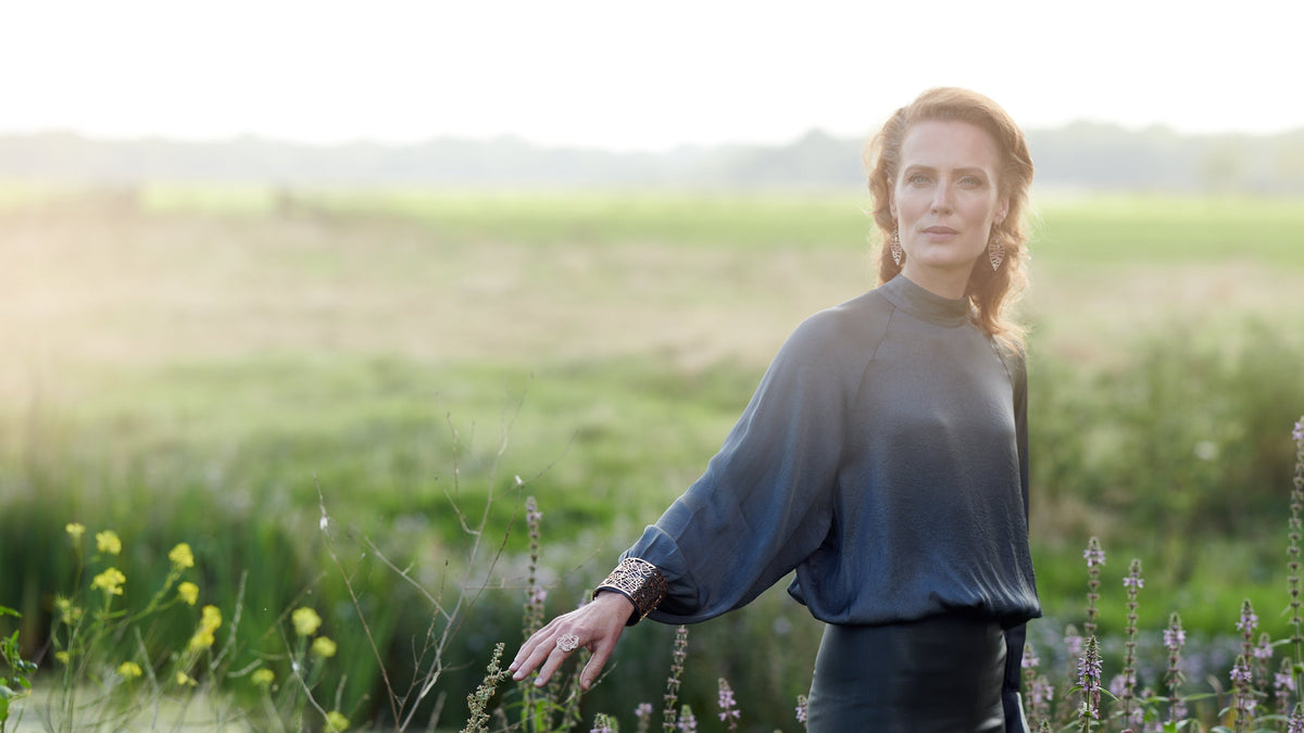 A woman stands in a sunlit field of wildflowers, wearing a De Parme Design rose gold cuff bracelet and ring in a leaf design.
