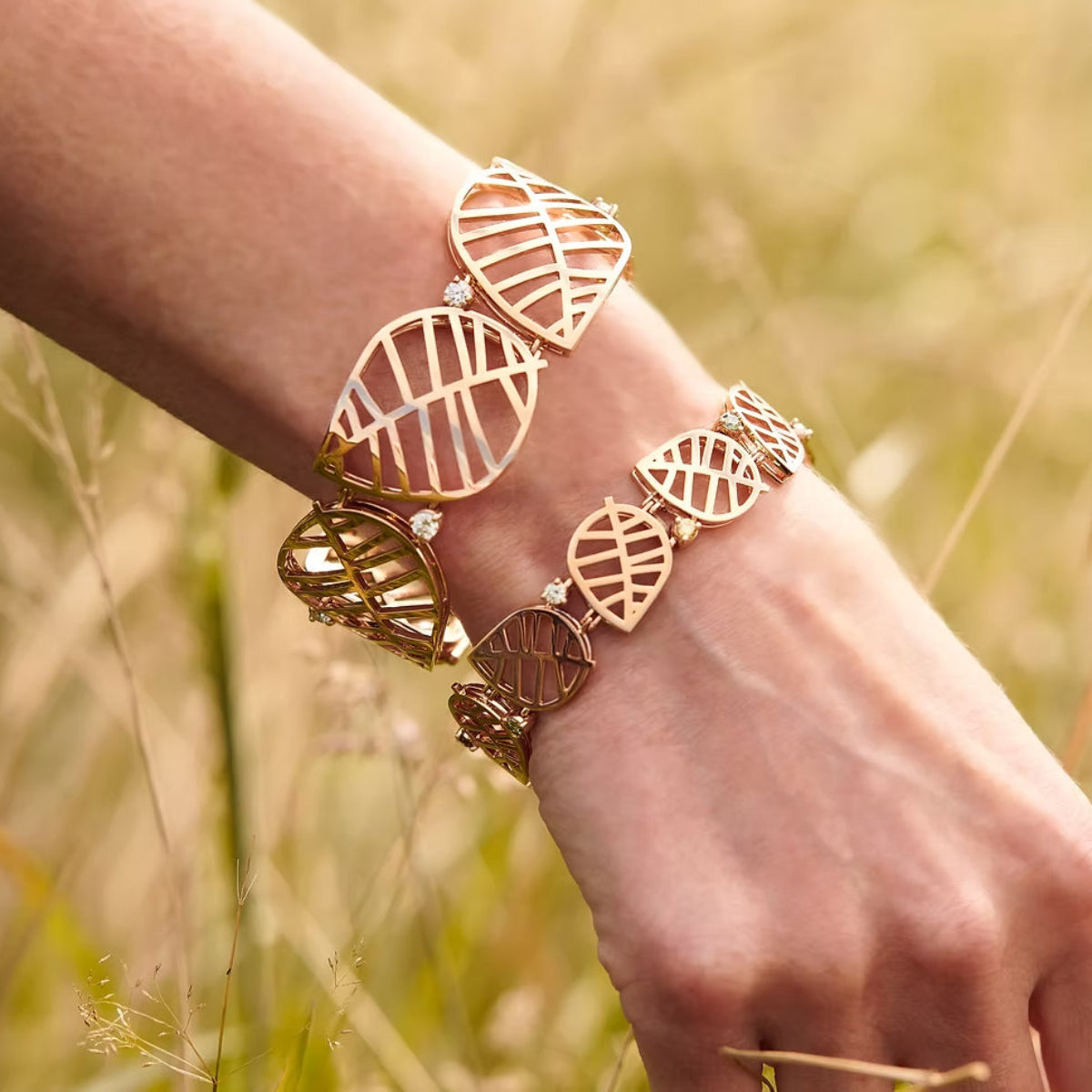 Two rose gold leaf-patterned cuff bracelets set with white diamonds, shown worn on a person's wrist against a blurred outdoor grass background.